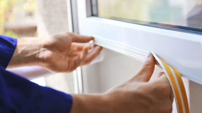Hands installing adhesive weatherstripping on a home window frame to prevent drafts and improve winter energy efficiency.