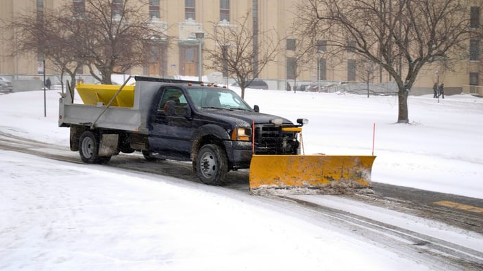 Commercial snow plow truck clearing snow on a commercial property during a winter storm, illustrating the importance of proactive snow removal planning and budgeting.