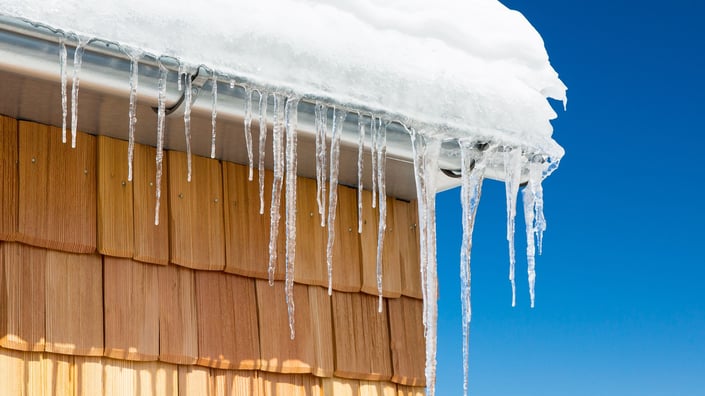ciIcicles hanging from the edge of a roof with snow buildup, illustrating the formation of ice dams on a residential home during winter.